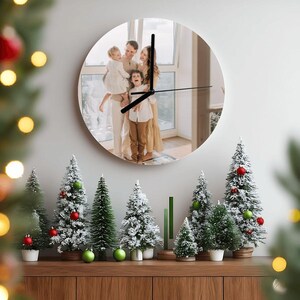 May include: A round wall clock with a family photo as the clock face. The clock hands are black. The clock is mounted on a white wall above a wooden cabinet decorated with miniature Christmas trees.