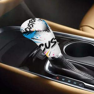 Könnte beinhalten: Ein individueller Schalthebelknauf mit weißer Basis, mit dem Wort "CUSTOM" in Schwarz, mit blauen und gelben Farbspritzern. Der Schalthebelknauf ist in einem Auto eingebaut.