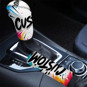 Könnte beinhalten: Ein weißer Schalthebelbezug für Autos mit dem Wort "CUSTOM" in schwarzer Graffiti-Schrift. Der Bezug hat farbenfrohe Farbspritzer in Blau, Pink, Gelb und Rot. Ein passender Armlehnenbezug ist ebenfalls sichtbar.