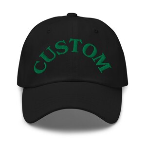 Puede incluir: Gorra de béisbol negra con la palabra "CUSTOM" bordada en verde en la parte delantera. El texto está arqueado. La gorra tiene una visera curva y un botón en la parte superior.