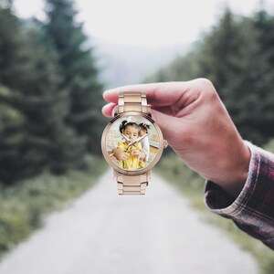 Puede incluir: Un reloj de pulsera de oro rosa con una esfera redonda que muestra una fotograf&iacute;a de un ni&ntilde;o. El reloj tiene una correa met&aacute;lica y una esfera de reloj anal&oacute;gica cl&aacute;sica con manecillas e indicadores de hora. El reloj se sostiene contra un fondo borroso al aire libre.