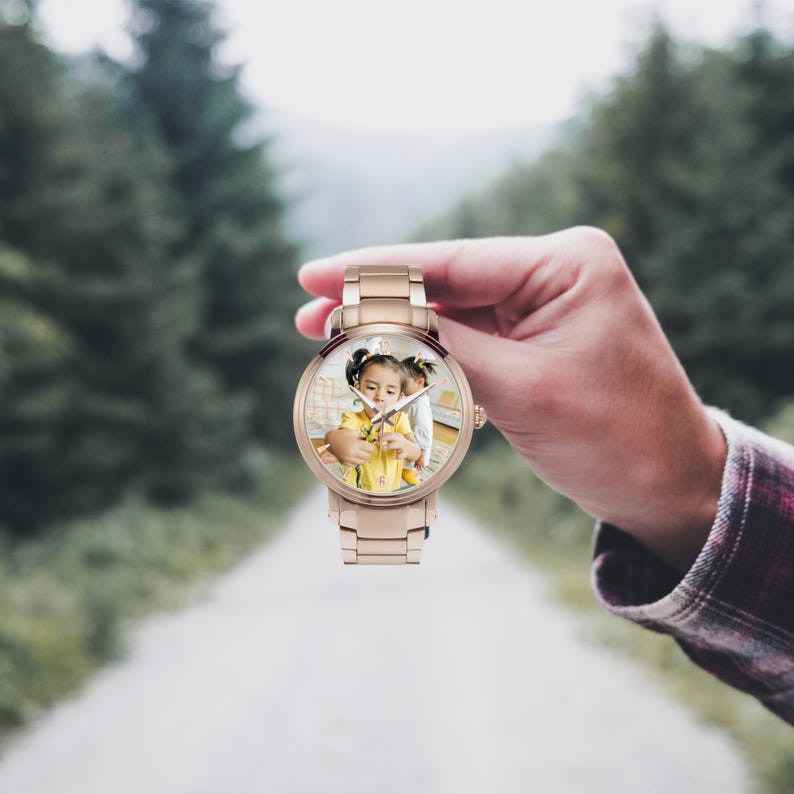Puede incluir: Un reloj de pulsera de oro rosa con una esfera redonda que muestra una fotograf&iacute;a de un ni&ntilde;o. El reloj tiene una correa met&aacute;lica y una esfera de reloj anal&oacute;gica cl&aacute;sica con manecillas e indicadores de hora. El reloj se sostiene contra un fondo borroso al aire libre.