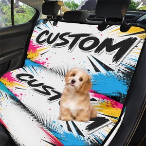 May include: Custom car seat cover with a colorful, abstract design featuring the word "CUSTOM" in bold black lettering. A small, light brown dog sits on the seat cover, adding a playful element to the design.