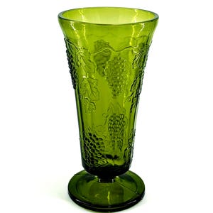 Op de afbeelding: Groene glazen vaas met een voetstuk. De vaas heeft een gedetailleerd druiven- en bladerontwerp. Het glas is licht transparant. De vaas is ongeveer 20 cm hoog.