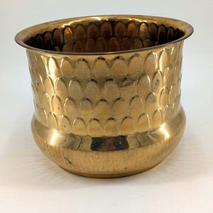 May include: A polished brass planter featuring a rounded base and a wide, flared rim. The exterior surface is decorated with a repeating embossed scale pattern that wraps around the upper portion of the vessel, while the lower section remains smooth.