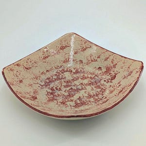 May include: A square ceramic bowl with a unique design. The bowl features a cream-colored base with a speckled red pattern. The rim is a darker red. The bowl is empty and sits on a white surface.