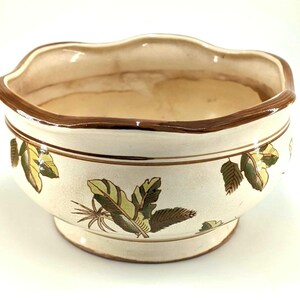 May include: A cream-colored ceramic bowl with a scalloped rim and a brown painted edge. The exterior features a repeating pattern of green and brown tropical leaves. The bowl has a circular base and a smooth, glossy finish throughout the piece.