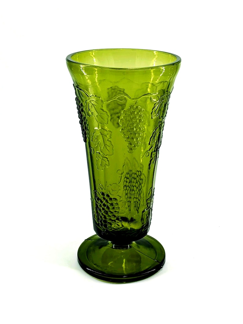 Op de afbeelding: Groene glazen vaas met een voetstuk. De vaas heeft een gedetailleerd druiven- en bladerontwerp. Het glas is licht transparant. De vaas is ongeveer 20 cm hoog.