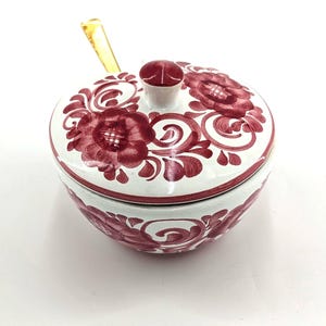 May include: A white ceramic sugar bowl with a lid featuring hand-painted dark red floral patterns and swirling leaf designs. The bowl includes a small matching lid with a central knob and a gold-toned metal spoon resting inside the container.