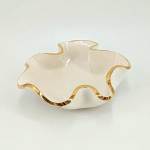 May include: A small, white ceramic dish with a ruffled edge and gold trim. The dish has a unique, organic shape and is likely used for serving small items or as a decorative piece. The gold trim adds a touch of elegance.
