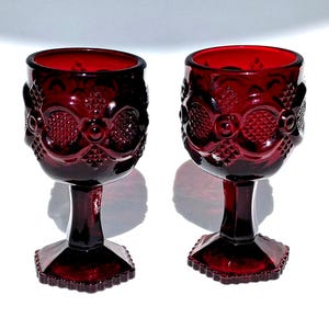 May include: Two ruby red goblets with ornate, embossed patterns. The stemware features a detailed design on the bowl and a faceted base. The glasses are made of a deep red glass, perfect for serving wine or other beverages.