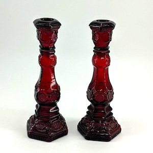 May include: Two ruby red glass candle holders. The candle holders have a detailed, ornate design with a square base and a flared top. The glass has a glossy finish, and the candle holders are identical in shape and size.
