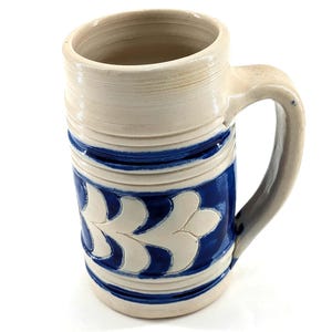 May include: A cylindrical ceramic beer mug featuring a light beige base color with a prominent cobalt blue band around the center. The blue section is decorated with a repeating white leaf-like pattern. The mug has a sturdy handle and a smooth rim.