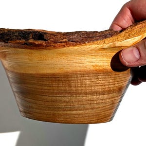 K&ouml;nnte beinhalten: Eine handgefertigte Holzschale mit einem nat&uuml;rlichen, rauen Rindenrand. Das Innere der Schale weist eine glatte, hellfarbige Holzmaserung auf, die an der Basis in ein dunkleres, sattes Braun &uuml;bergeht. Die Schale wird von einer Hand gehalten, die ihre organische Form zeigt.