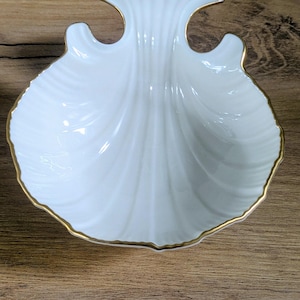 May include: A white, shell-shaped dish with a gold trim. The dish has a detailed, ridged design and is set against a wooden surface. The dish is likely made of porcelain or ceramic and is a decorative item.