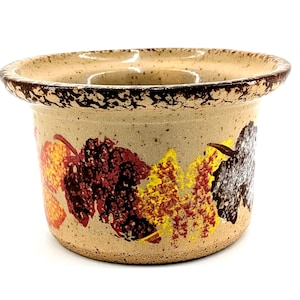 May include: A ceramic candle holder with a speckled beige finish and a dark brown rim. The holder is decorated with colorful leaf designs in shades of red, yellow, and brown. The holder has a circular opening.