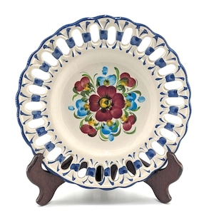 May include: A round ceramic plate with a cream-colored center featuring a hand-painted floral motif in red, blue, and green. The rim has a scalloped edge with a lattice cutout design, accented by blue painted details along the border.