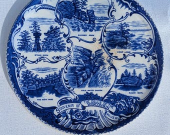 May include: A round, blue and white souvenir plate featuring scenic illustrations of the Wisconsin River Dells. The plate includes text such as "Chimney Rock", "Stand Rock", and "Sugar Bowl". The border has a decorative pattern.