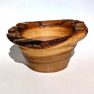 K&ouml;nnte beinhalten: Eine handgefertigte Holzschale mit einem nat&uuml;rlichen, rauen Rand. Die Schale hat eine hellbraune und beige Farbgebung mit sichtbarer Holzmaserung. Die Au&szlig;enseite hat eine ausgestellte Form und der Rand eine dunkle, strukturierte Rinde.