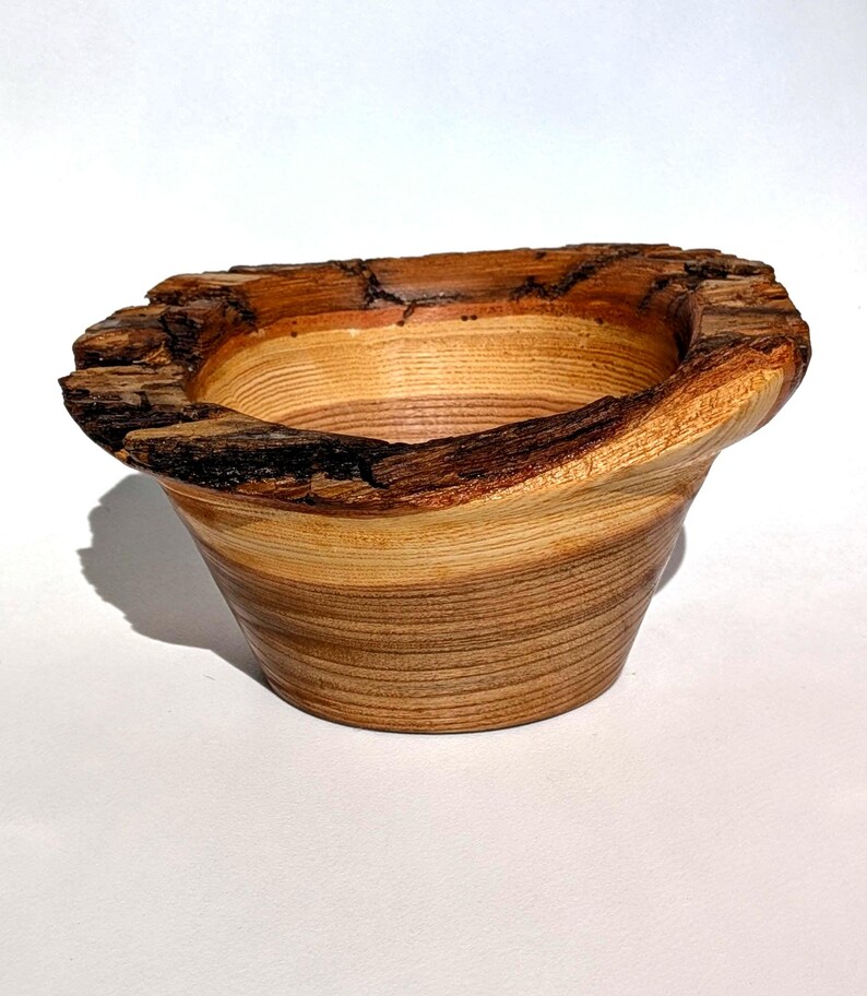 K&ouml;nnte beinhalten: Eine handgefertigte Holzschale mit einem nat&uuml;rlichen, rauen Rand. Die Schale hat eine hellbraune und beige Farbgebung mit sichtbarer Holzmaserung. Die Au&szlig;enseite hat eine ausgestellte Form und der Rand eine dunkle, strukturierte Rinde.