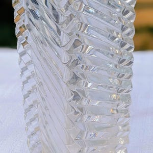 May include: Clear glass vase with a textured, geometric design. The vase has a rectangular shape with a series of diagonal, ridged patterns that create a spiral effect. The glass reflects light, creating a sparkling appearance.