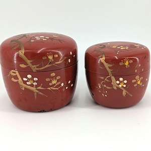 May include: Two antique, red lacquered boxes with lids. The boxes are decorated with gold and white floral designs. The boxes are oval-shaped and have a glossy finish. The boxes are likely used for storage or display.