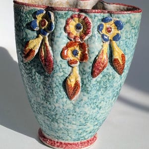 May include: A turquoise ceramic vase with a textured surface and a red rim. The vase is adorned with three-dimensional floral designs in yellow, red, and blue. The base of the vase also has a red trim.