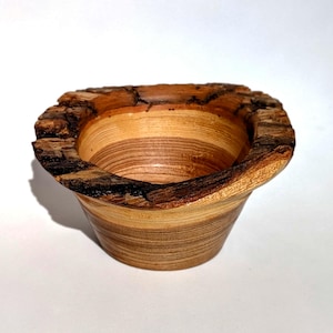 K&ouml;nnte beinhalten: Eine handgefertigte Holzschale mit einem nat&uuml;rlichen, rauen Rand. Die Schale hat ein hellbraunes und beigefarbenes Streifenmuster, das die Maserung des Holzes zeigt. Die Au&szlig;enseite ist glatt, w&auml;hrend der Rand seine rohe, rindenartige Textur beh&auml;lt.