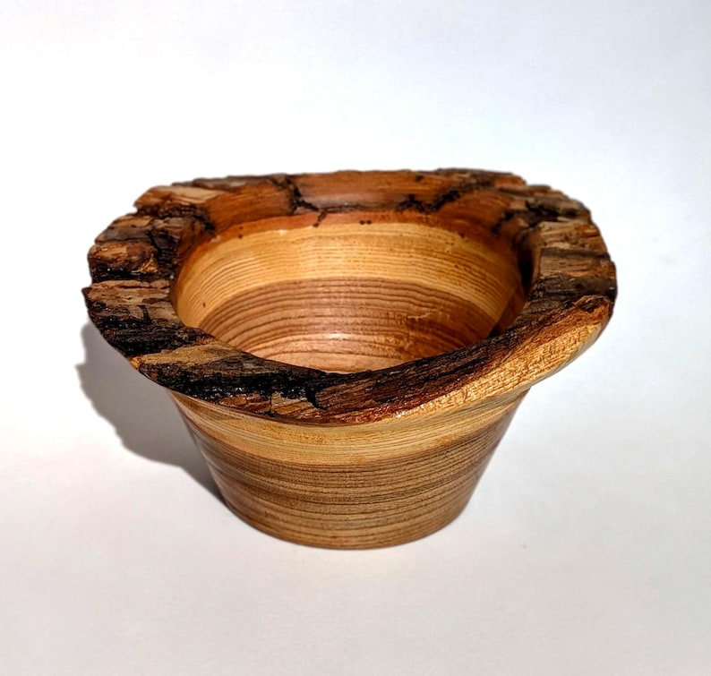 K&ouml;nnte beinhalten: Eine handgefertigte Holzschale mit einem nat&uuml;rlichen, rauen Rand. Die Schale hat ein hellbraunes und beigefarbenes Streifenmuster, das die Maserung des Holzes zeigt. Die Au&szlig;enseite ist glatt, w&auml;hrend der Rand seine rohe, rindenartige Textur beh&auml;lt.