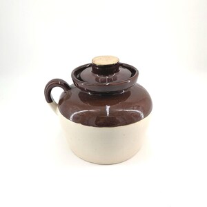 May include: A ceramic crock with a rounded body, featuring a glossy dark brown glaze on the upper portion and a matte cream-colored base. It includes a matching dark brown lid with a central knob and a single curved handle on the side.