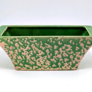 May include: A rectangular ceramic planter with a green interior and exterior. The exterior has a textured, speckled pattern in a light pink color. The planter has a trapezoidal shape and is suitable for indoor or outdoor use.