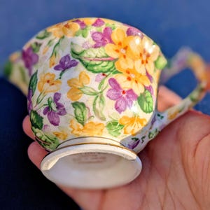 May include: A small, floral-patterned creamer with a handle. The creamer features a white base and is adorned with yellow, purple, and green floral designs. The creamer is held in a hand.