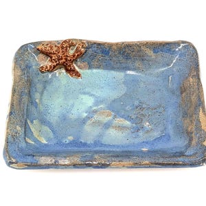 May include: A handmade ceramic tray with a blue and brown glaze. A brown starfish is attached to the upper left corner. The tray is rectangular with slightly uneven edges, giving it a rustic appearance.