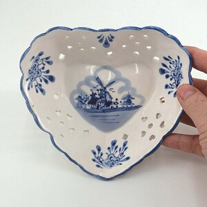 May include: A heart-shaped ceramic bowl with a white base and blue painted details. The center features a blue illustration of a windmill. The rim is scalloped with blue edges and includes a series of small, cut-out diamond-shaped perforations.