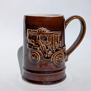 May include: A brown ceramic mug with a handle, featuring a detailed relief of a vintage car with passengers. The mug has a glossy finish and a cream-colored interior. The car's design includes wheels, a roof, and figures.
