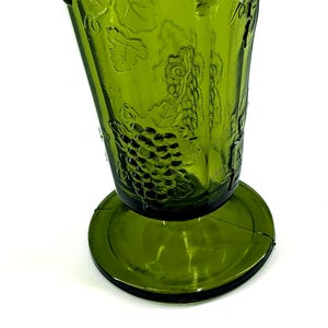 Op de afbeelding: Een groene glazen beker met een brede basis en een uitlopende bovenkant. De beker heeft een reli&euml;f druiven- en wijnrankenontwerp. Het glas heeft een lichte tint en staat tegen een witte achtergrond. De beker is waarschijnlijk vintage.