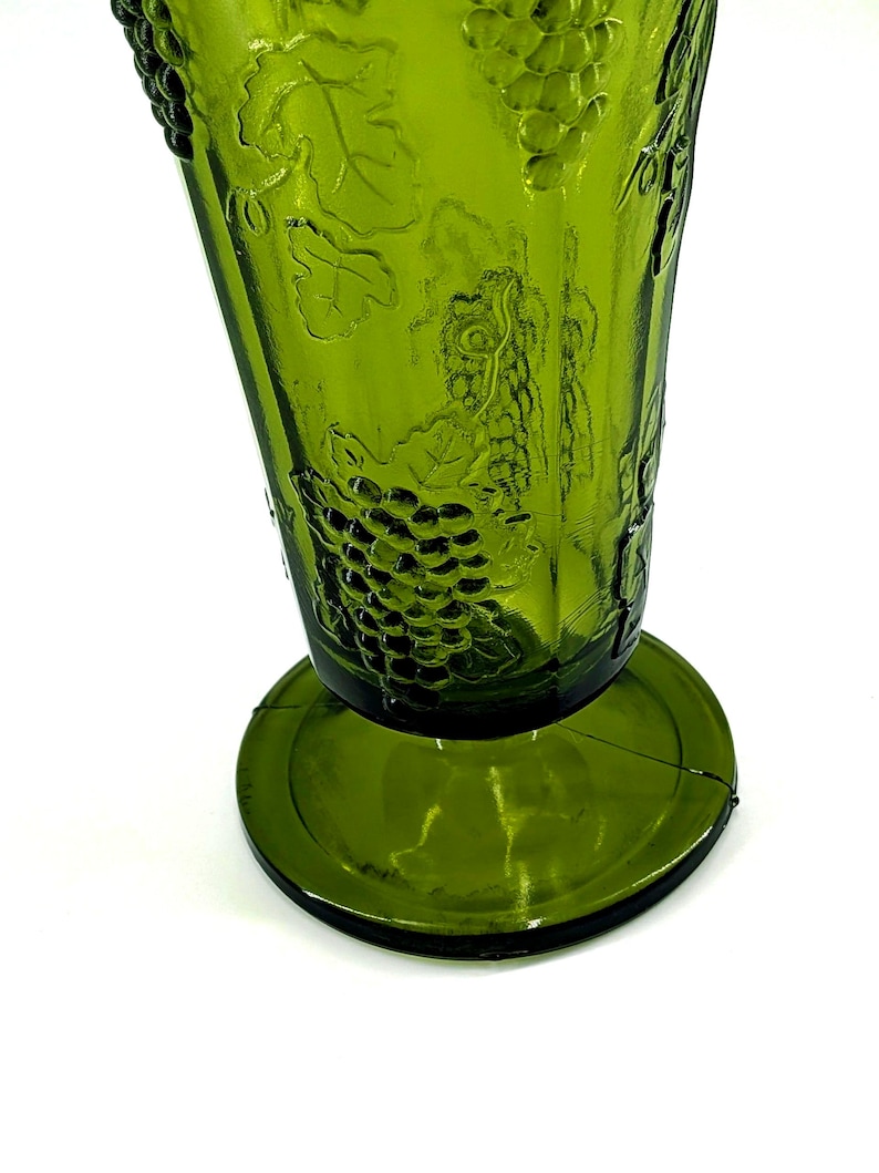 Op de afbeelding: Een groene glazen beker met een brede basis en een uitlopende bovenkant. De beker heeft een reli&euml;f druiven- en wijnrankenontwerp. Het glas heeft een lichte tint en staat tegen een witte achtergrond. De beker is waarschijnlijk vintage.