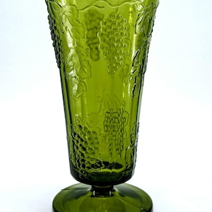 Op de afbeelding: Groene glazen vaas met een uitlopende bovenkant en een ronde voet. De vaas heeft een reli&euml;fontwerp van druiven en bladeren. Het glas heeft een glanzende afwerking en is effen groen van kleur. De vaas is waarschijnlijk vintage en geschikt voor bloemstukken.