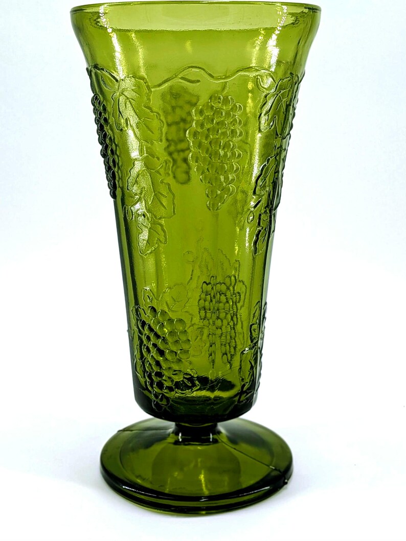 Op de afbeelding: Groene glazen vaas met een uitlopende bovenkant en een ronde voet. De vaas heeft een reli&euml;fontwerp van druiven en bladeren. Het glas heeft een glanzende afwerking en is effen groen van kleur. De vaas is waarschijnlijk vintage en geschikt voor bloemstukken.