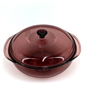 May include: A vintage, round, purple glass casserole dish with a matching lid. The lid has a central knob and a ridged design. The dish has two handles and is designed for oven use. The glass is a deep, translucent purple.