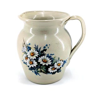 May include: A beige ceramic pitcher with a handle and a wide spout. The pitcher is decorated with a floral design featuring white daisies with yellow centers and blue accents. The pitcher is a decorative item.