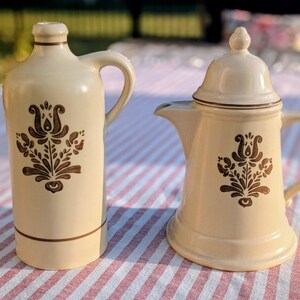 May include: Two cream-colored ceramic kitchenware items. One is a tall, handled bottle with a brown floral design. The other is a lidded coffee pot with a spout and a matching floral pattern. Both items have brown trim.