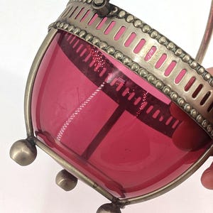 May include: A vintage glass basket with a vibrant pink hue. The basket features a silver-toned metal frame with a handle and decorative detailing. The base has three rounded feet.