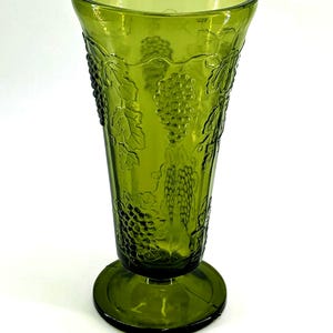 Op de afbeelding: Groene glazen vaas met een voetstuk. De vaas heeft een reli&euml;f druiven- en bladerontwerp. De vaas heeft een uitlopende bovenkant en is ongeveer 20 cm hoog.