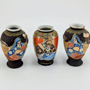May include: Three small ceramic vases with a matte black finish. Each vase features hand-painted figures in colorful traditional attire, gold-tone accents, and intricate patterns around the neck and base. The central vase has an orange panel.