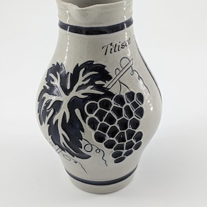 May include: A gray ceramic pitcher with a dark blue grape and leaf design. The word "Titisee" is written in cursive. The pitcher has a dark blue band around the top and bottom.