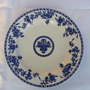 May include: A white ceramic plate with a blue floral design. The plate features a central motif of a basket of flowers within a circle, surrounded by a border of flowers and leaves. The rim has a blue patterned edge.