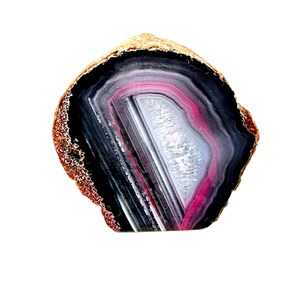 May include: A polished agate stone slice with concentric bands of black, white, and pink. The outer edge has a rough, golden-brown texture. The stone is cut to reveal the natural patterns and colors.