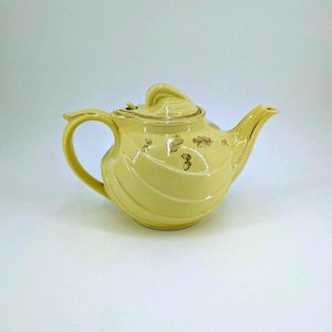 May include: A pale yellow ceramic teapot with a curved handle and spout. The teapot has a lid and is decorated with gold-colored leaf and acorn designs. The rim and handle are also trimmed in gold. The teapot is a vintage style.