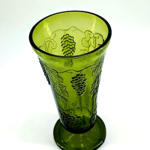 Op de afbeelding: Een groene glazen vaas met een uitlopende bovenkant en een ronde basis. De vaas is versierd met reli&euml;f druiven- en bladerenontwerpen. Het glas heeft een lichte groene tint en de vaas is waarschijnlijk vintage.